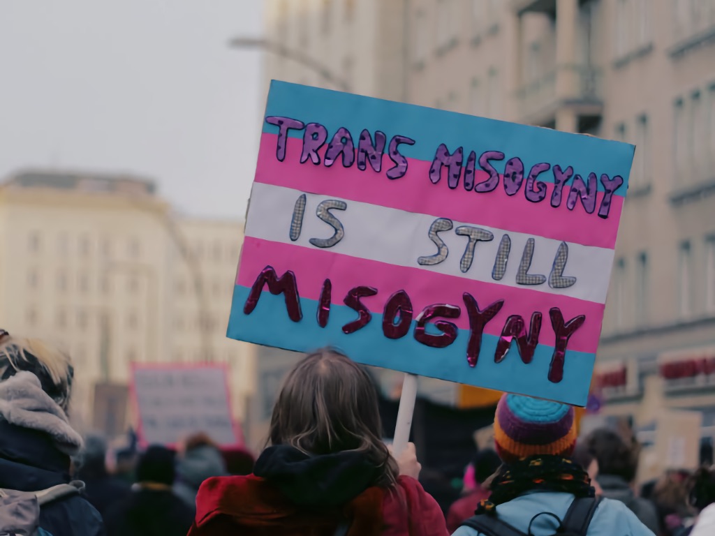 A photo taken at a trans rights protest. It shows a protestor with their back to the camera, holding a sign in the signature trans flag colours which reads "Trans Misogyny is still Misogyny".